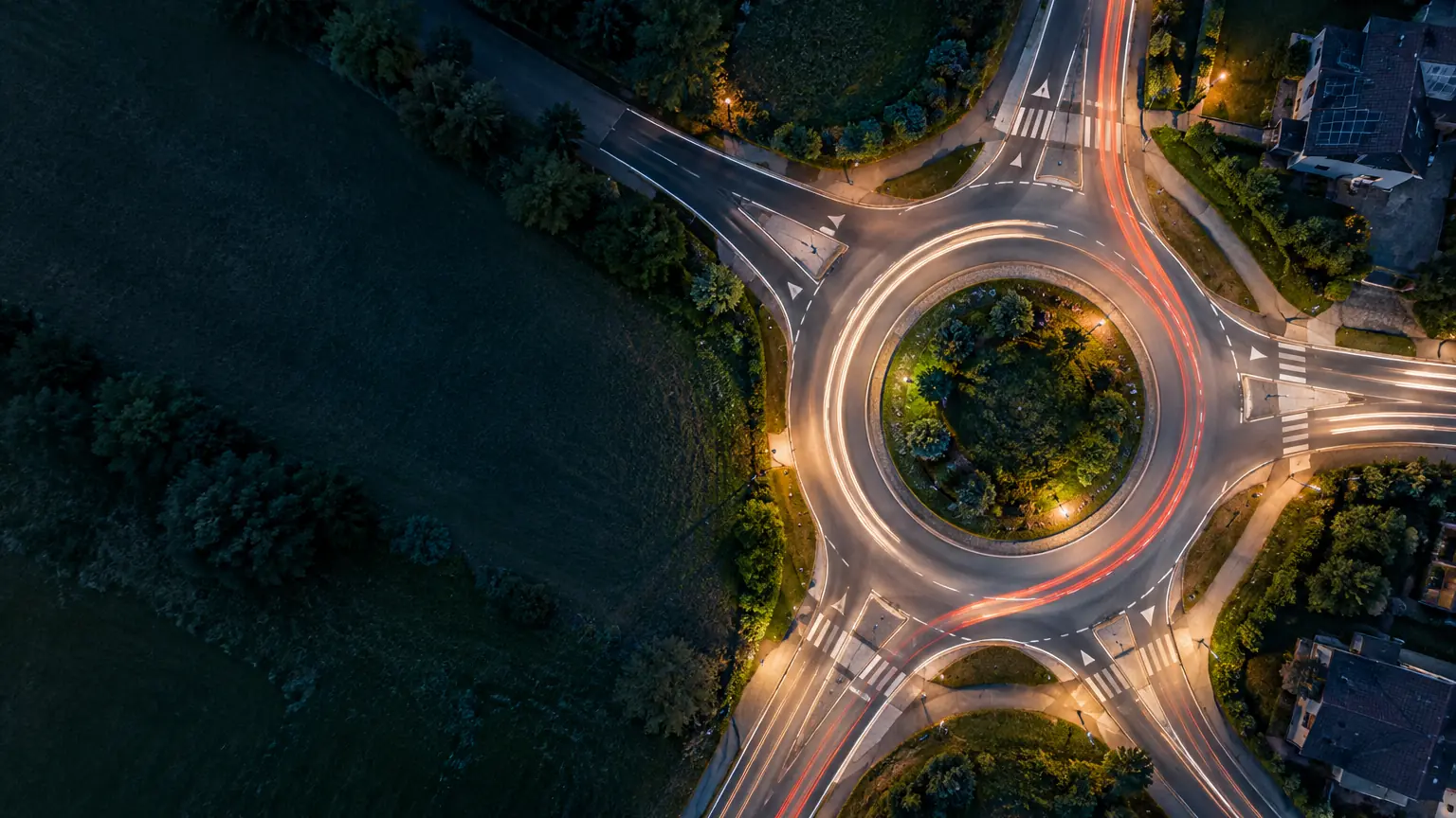Aerial view of a roundabout at dusk. Vehicle light trails circle a central island with trees and shrubs. A dark grassy field lies to the left, with a nearly unlit road leading towards the upper left.
